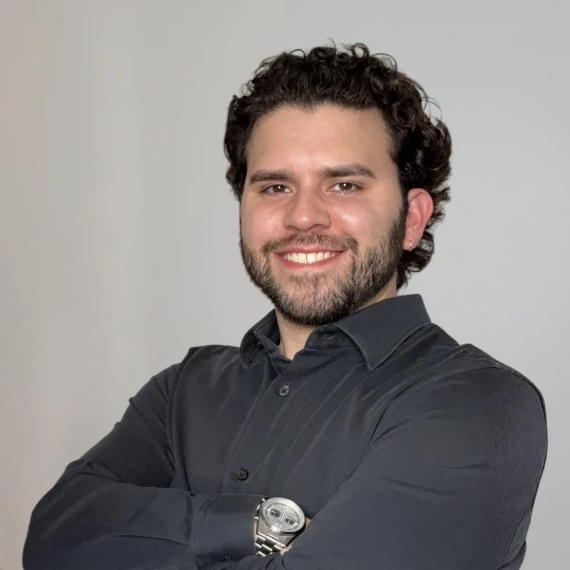 A smiling man with curly hair wearing a black shirt and a wristwatch, posing with crossed arms against a plain background.