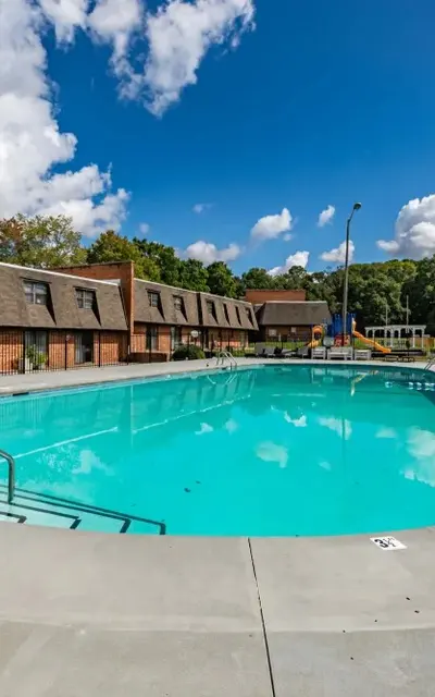 A clear blue swimming pool surrounded by a concrete deck and a fenced area. Nearby, there are several apartment buildings with sloped roofs and trees in the background under a partly cloudy sky.