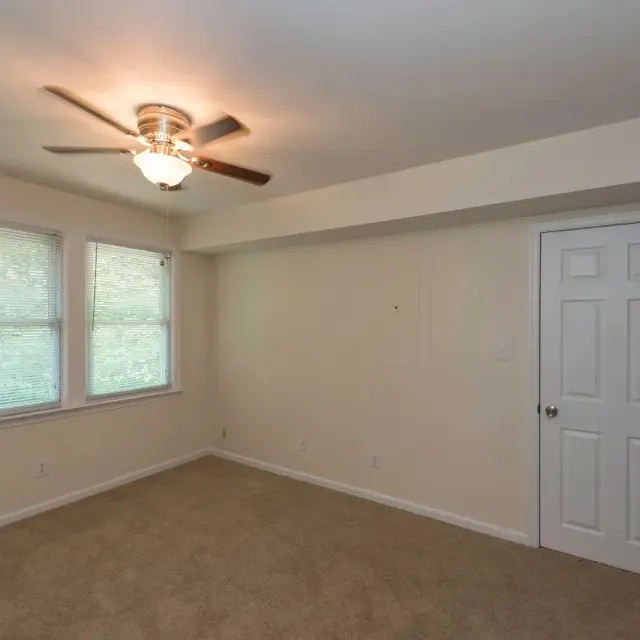 A spacious empty room with beige carpet flooring, a ceiling fan, and two windows allowing natural light to enter. There is a closed door on the right side leading to another space.