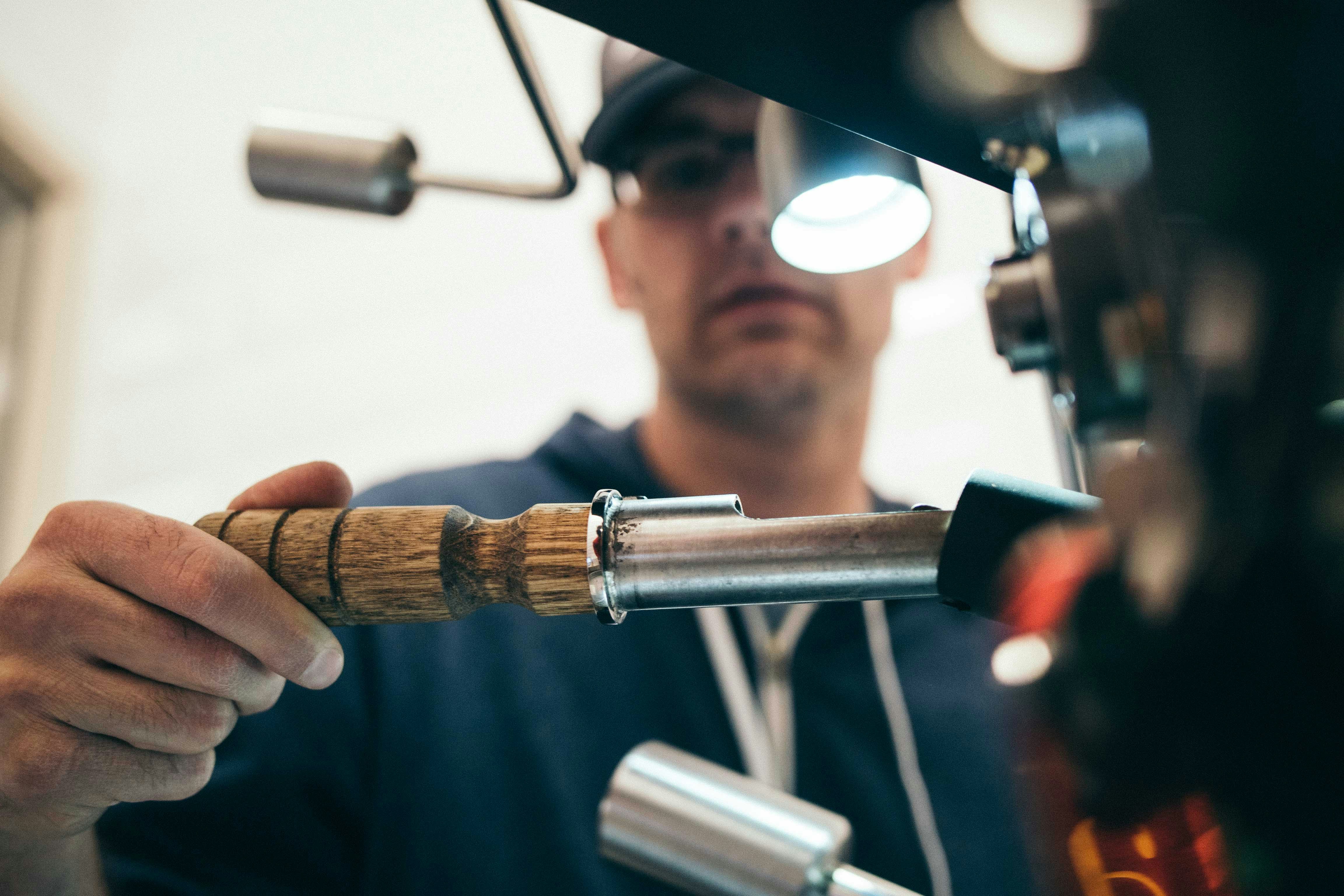 A barista carefully operates an espresso machine, holding a metallic tool with a wooden handle. A focused expression is visible on their face as they work under a bright light.
