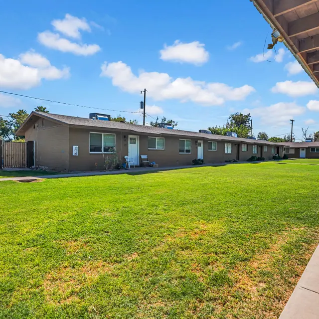 View of a low-rise apartment complex from a sidewalk, featuring multiple units and a large green lawn.