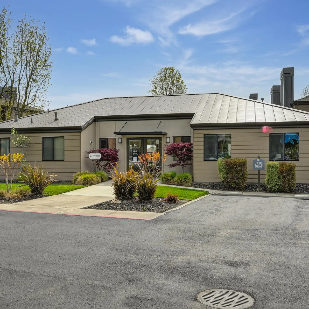 A modern apartment complex features a single-story building with landscaped gardens, shrubs, and small trees.