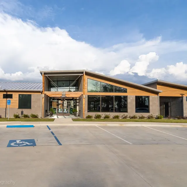 A modern building with a large glass front, wooden accents, and a parking area in front under a blue sky with some clouds.
