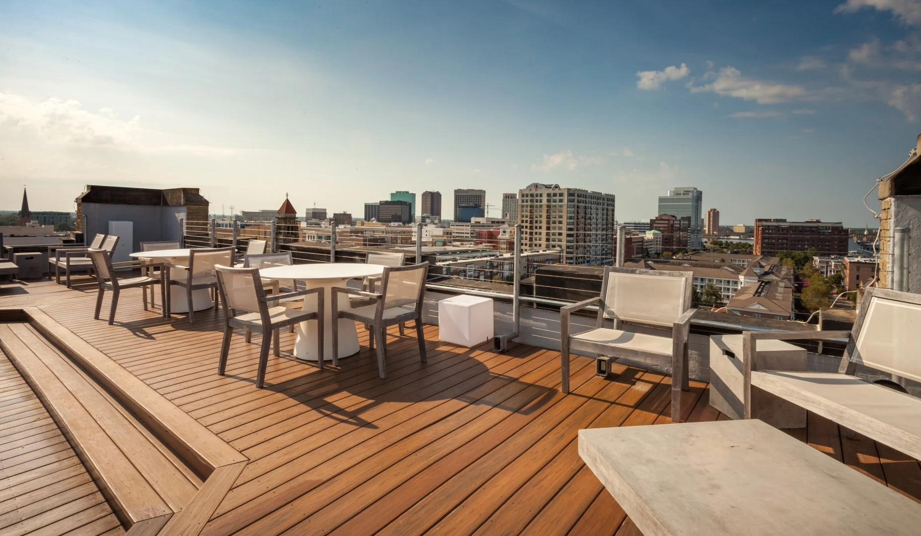 View from a rooftop terrace featuring wooden flooring, modern outdoor furniture, and a skyline of urban buildings in the background under a clear sky.