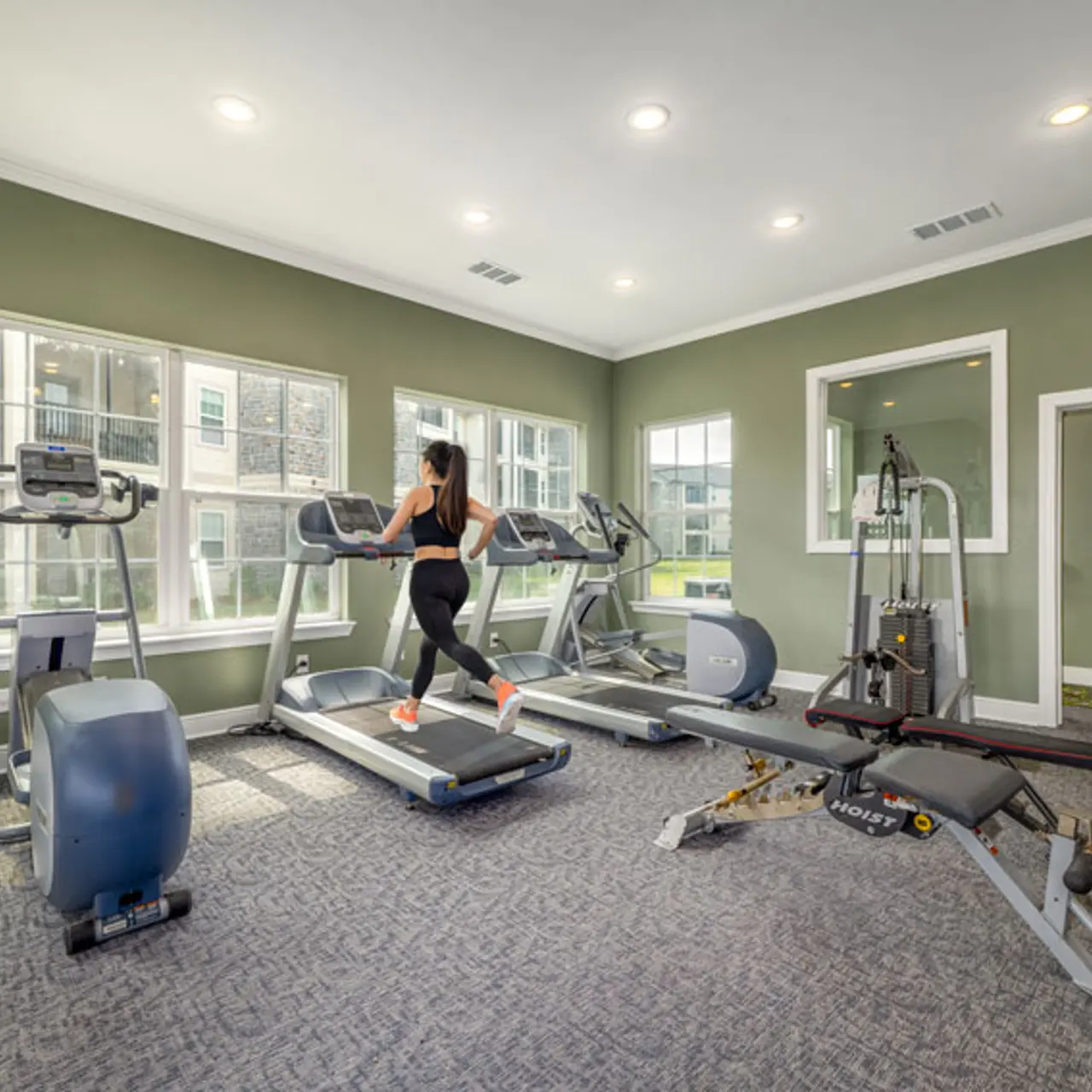 A person running on a treadmill in a modern gym with large windows, green walls, and various fitness equipment.