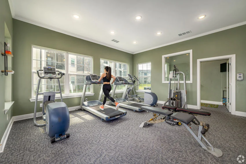 A person running on a treadmill in a modern gym with large windows, green walls, and various fitness equipment.
