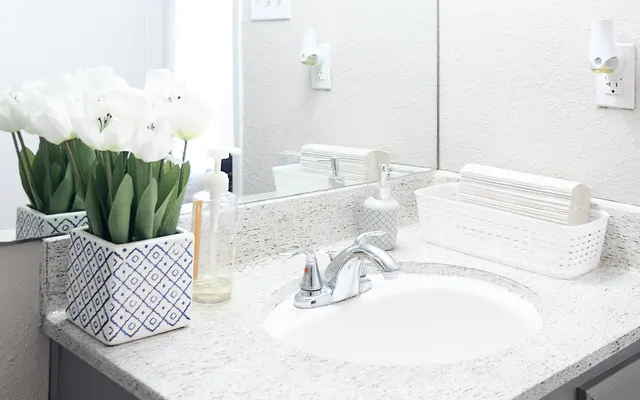 A modern bathroom sink area with a granite countertop, featuring a vase of white tulips, a soap dispenser, and a storage basket. The decor is minimalist with a mirror reflecting the light from a doorway.