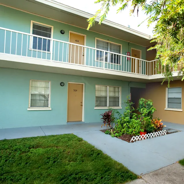 A two-story apartment building with a pastel color scheme featuring green and blue walls. The ground floor has two doors and a small landscaped area with plants and flowers. The upper floor has a balcony with railings.