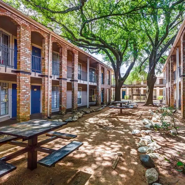 Apartment Complex Courtyard A courtyard of a brick apartment complex featuring two-story buildings and large trees providing shade. Picnic tables are arranged on a gravel surface with well-kept greenery around.