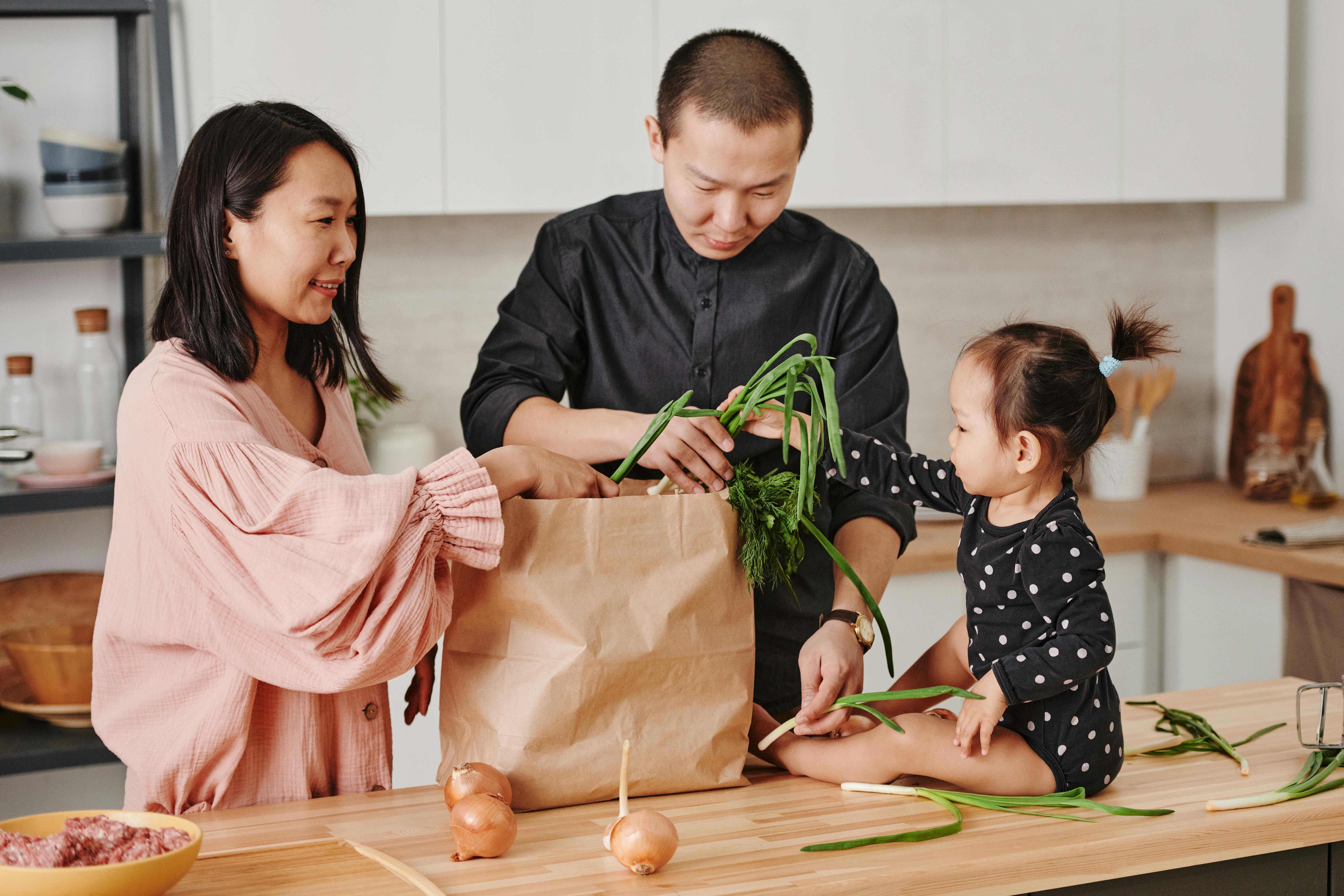 A family in a kitchen organizing ingredients from a brown paper bag. The woman on the left is wearing a pink sweater and is reaching into the bag, while a man in black is pulling out green onions. A young girl, sitting on the table, is holding some green onions and looking at her parents.