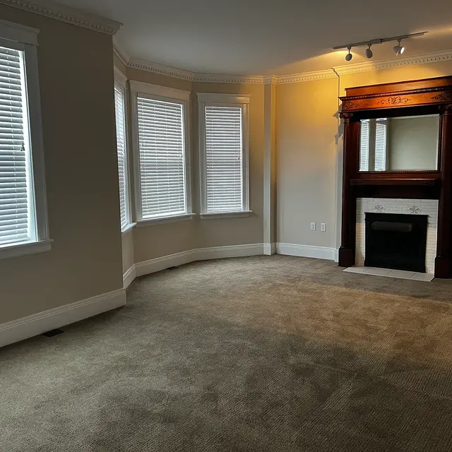 A spacious empty living room featuring large windows with blinds, a light beige wall color, and a decorative fireplace mantel against one wall.
