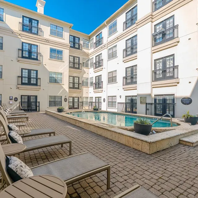A view of a well-maintained outdoor pool area surrounded by modern apartment buildings. Lounge chairs are arranged around the pool, with planters and a clear blue sky above.