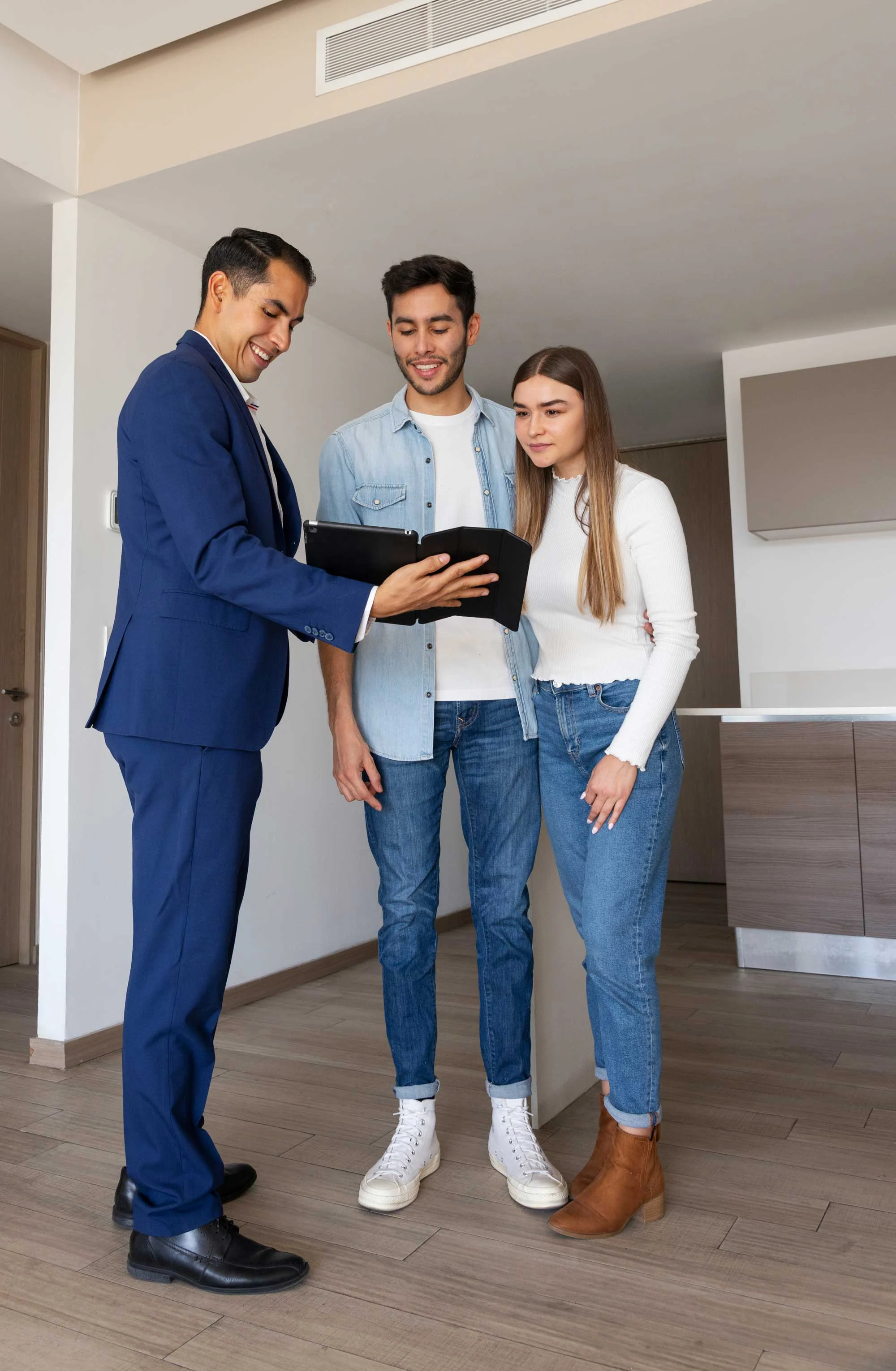 A real estate agent showing a laptop screen to a young couple who are standing in a modern, bright apartment. The agent is dressed in a suit, while the couple is casually dressed, looking engaged in the discussion.