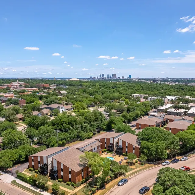 Aerial view of a suburban area featuring green trees, residential buildings, and a distant city skyline under a clear blue sky.