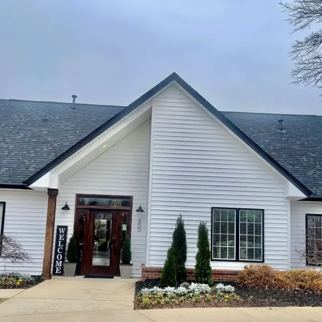 A modern welcoming building with a sloped roof, featuring white wooden siding and dark trim, surrounded by neatly landscaped plants and flowers.