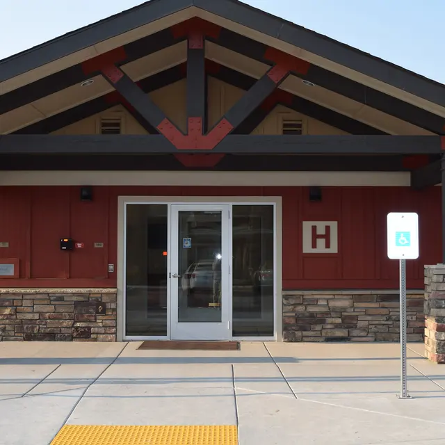 Building Entrance with Accessibility Features Front entrance of a building with a red wooden exterior, stone pillars, and double glass doors. The letter 'H' is displayed above the doors, and there are marked parking spaces nearby.