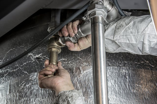 Hands working on plumbing, connecting metal pipes and fittings under a surface.