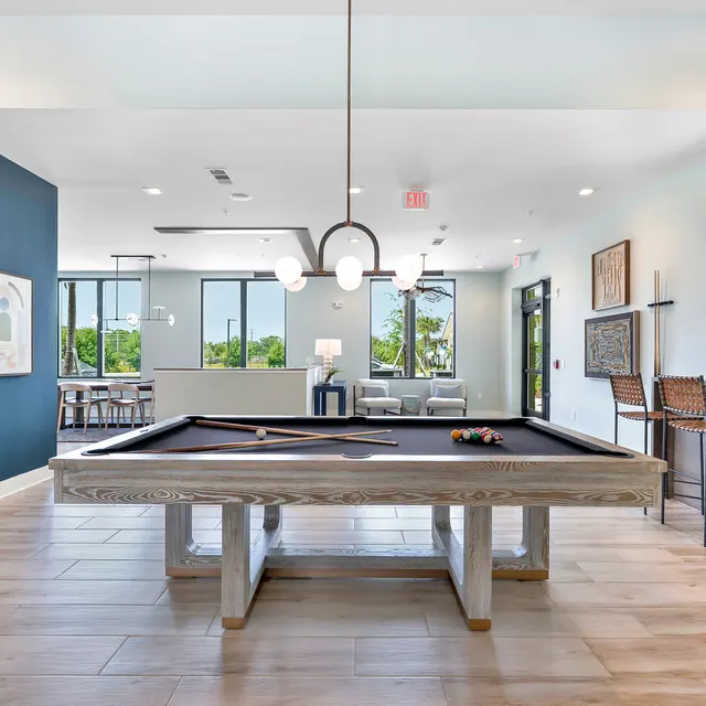 Modern Game Room with Pool Table A spacious game room featuring a pool table, modern furnishings, and large windows letting in natural light. The decor includes blue accent walls and wooden flooring.