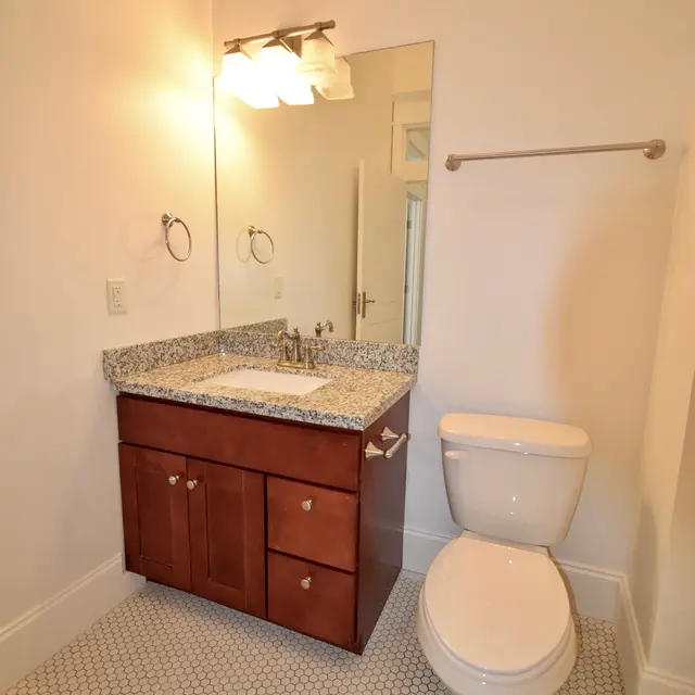 A modern bathroom featuring a wooden vanity with a granite countertop, a round mirror, and a toilet. The flooring consists of small white hexagonal tiles, complemented by bright lighting.