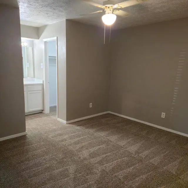 Empty Room with Ceiling Fan and Bathroom Access A clean, empty room with a ceiling fan and light fixture, featuring a beige carpet, and a bathroom door in the background.