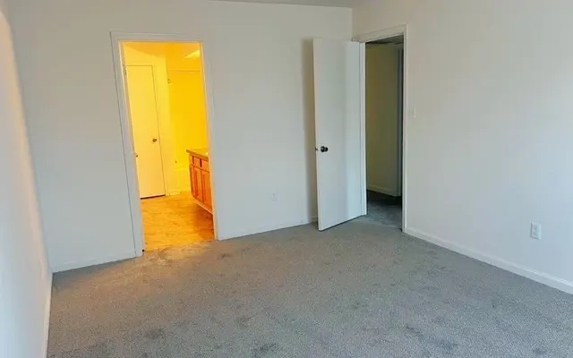 Empty Room Interior An empty room with beige carpet and lightly-colored walls. Two doors are visible: one leading to a bathroom area with warm lighting and another leading to a hallway or another room. The space appears well-lit and clean.