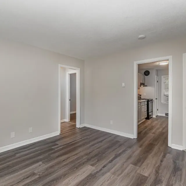 A spacious empty room with light grey walls and a hardwood floor, featuring doorways leading to other areas.