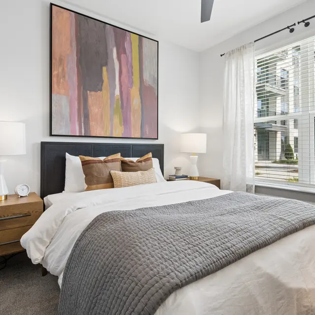 Stylish Modern Bedroom A well-decorated bedroom featuring a large bed with a gray throw blanket, two bedside tables with lamps, and a large window revealing a glimpse of an exterior view.