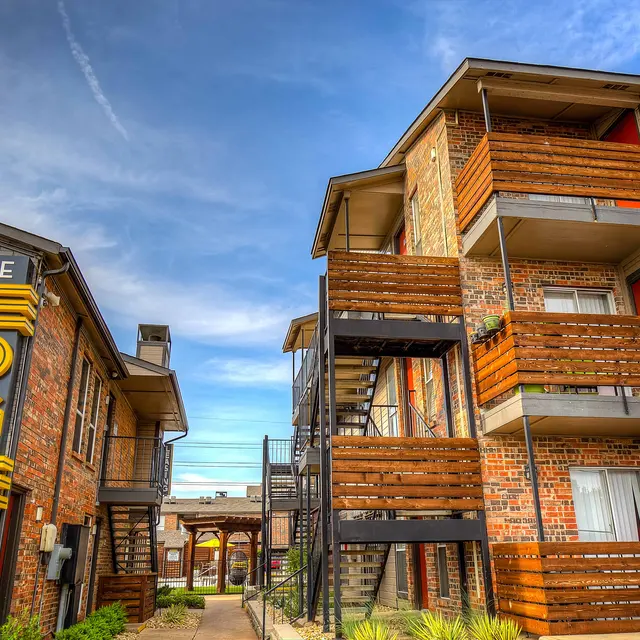 A two-building apartment complex named 'The Edge,' featuring modern architecture with exposed brick and wooden balconies. A clear blue sky is seen above.