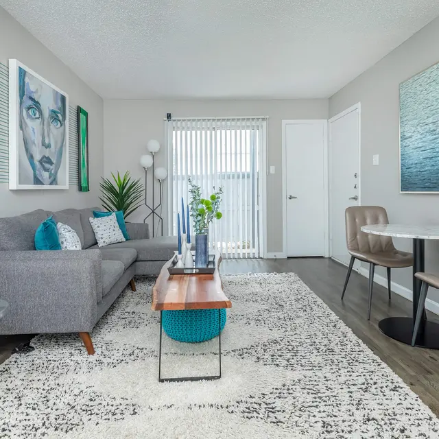 A modern living room with a gray sofa, a round wooden coffee table, and a vibrant abstract artwork. A rug covers the floor, and there are decorative plants and wall art in teal and turquoise colors.