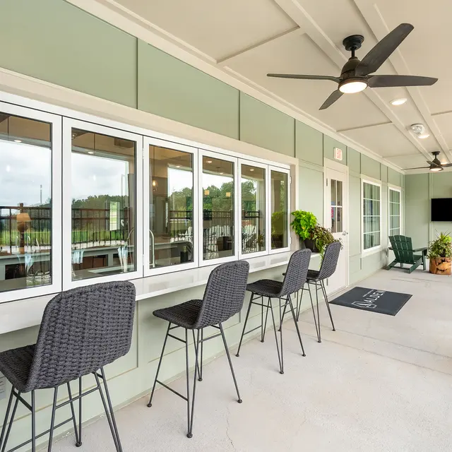 A spacious outdoor seating area with black chairs near large windows, offering a view of greenery. There are indoor plants and a TV mounted on the wall, with ceiling fans for ventilation.