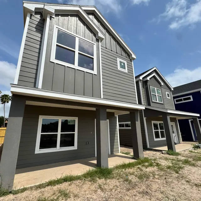 A row of newly constructed houses with a modern design. The house in the foreground features gray siding with white trim and a spacious porch. The background shows additional houses in blue and gray tones.