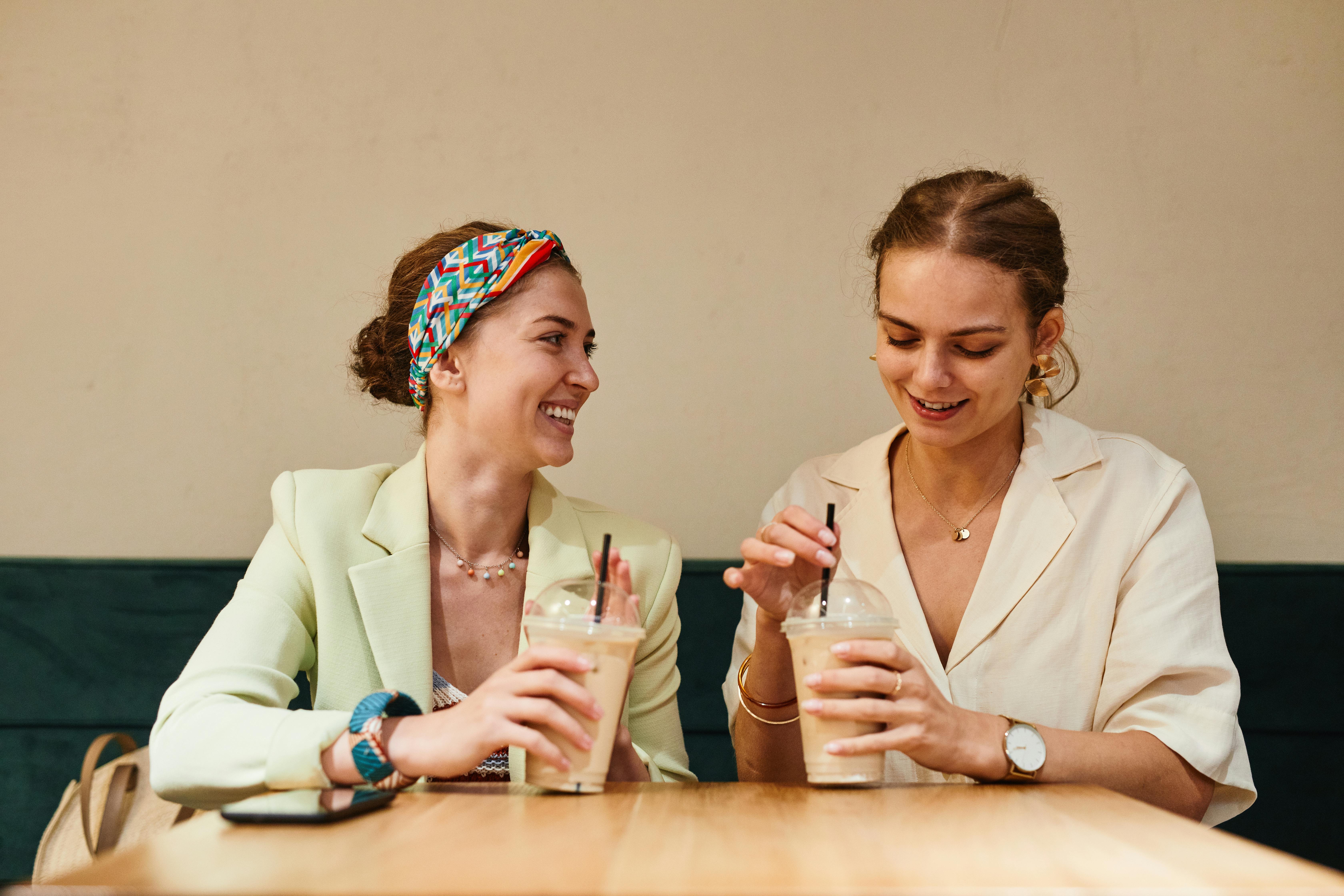Two women sitting at a table in a café, enjoying iced drinks and smiling at each other.