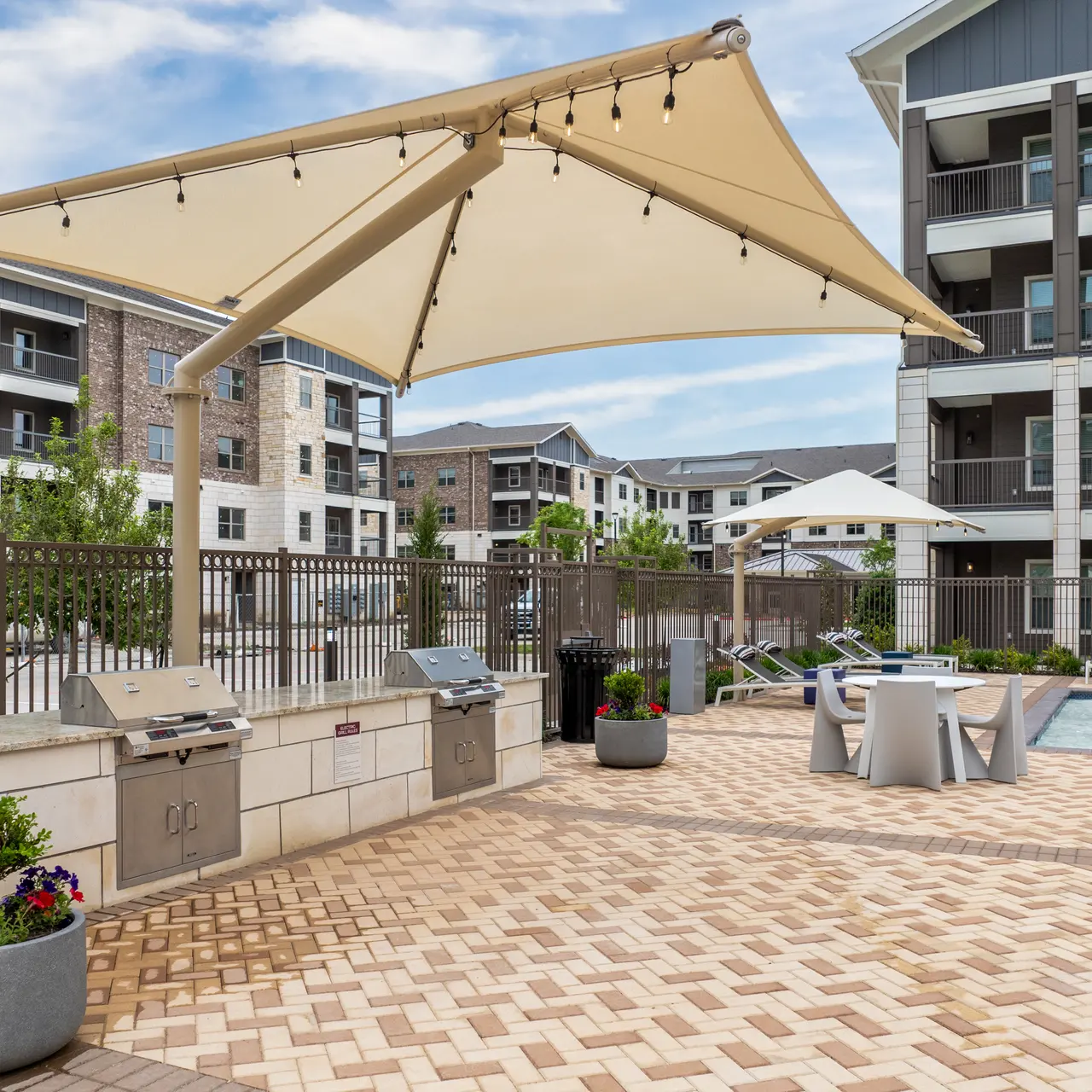 An outdoor area featuring a pool, shaded seating, and grill stations, surrounded by modern apartment buildings.