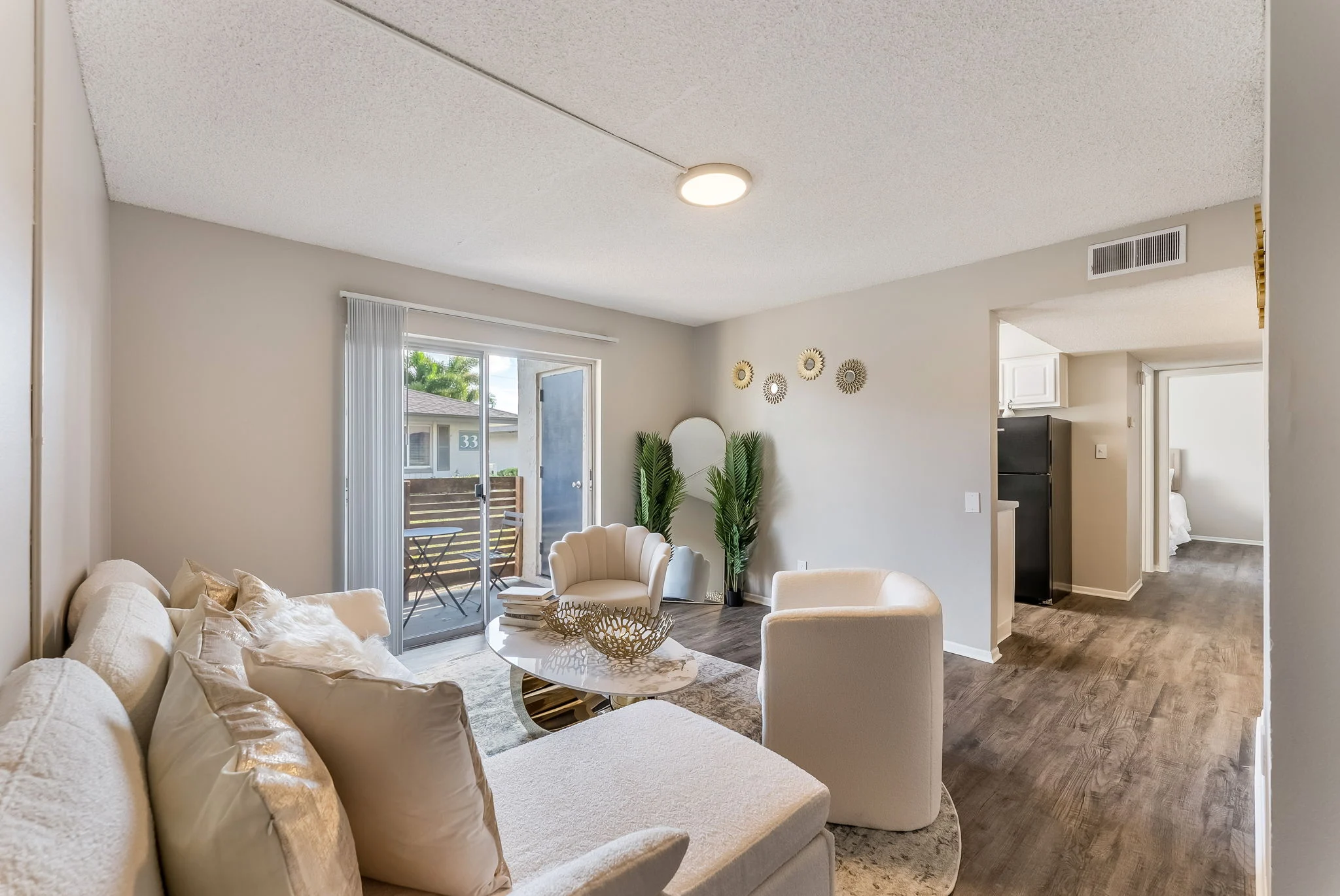 A modern and cozy living room featuring a light-colored sofa, decorative pillows, and a round coffee table. There are plants and a balcony view visible through sliding glass doors. The flooring is wood-like, and the walls are light-colored with decorative wall items.