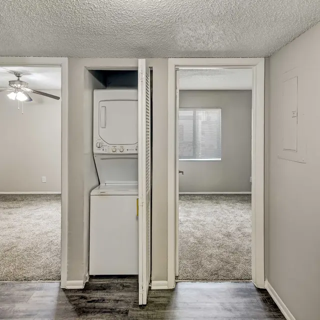 View of an apartment interior featuring a laundry area in the foreground with a washing machine and dryer, and two doorways leading to separate rooms. One room appears to have a window with natural light coming in, while the other room has a ceiling fan and carpeted flooring.
