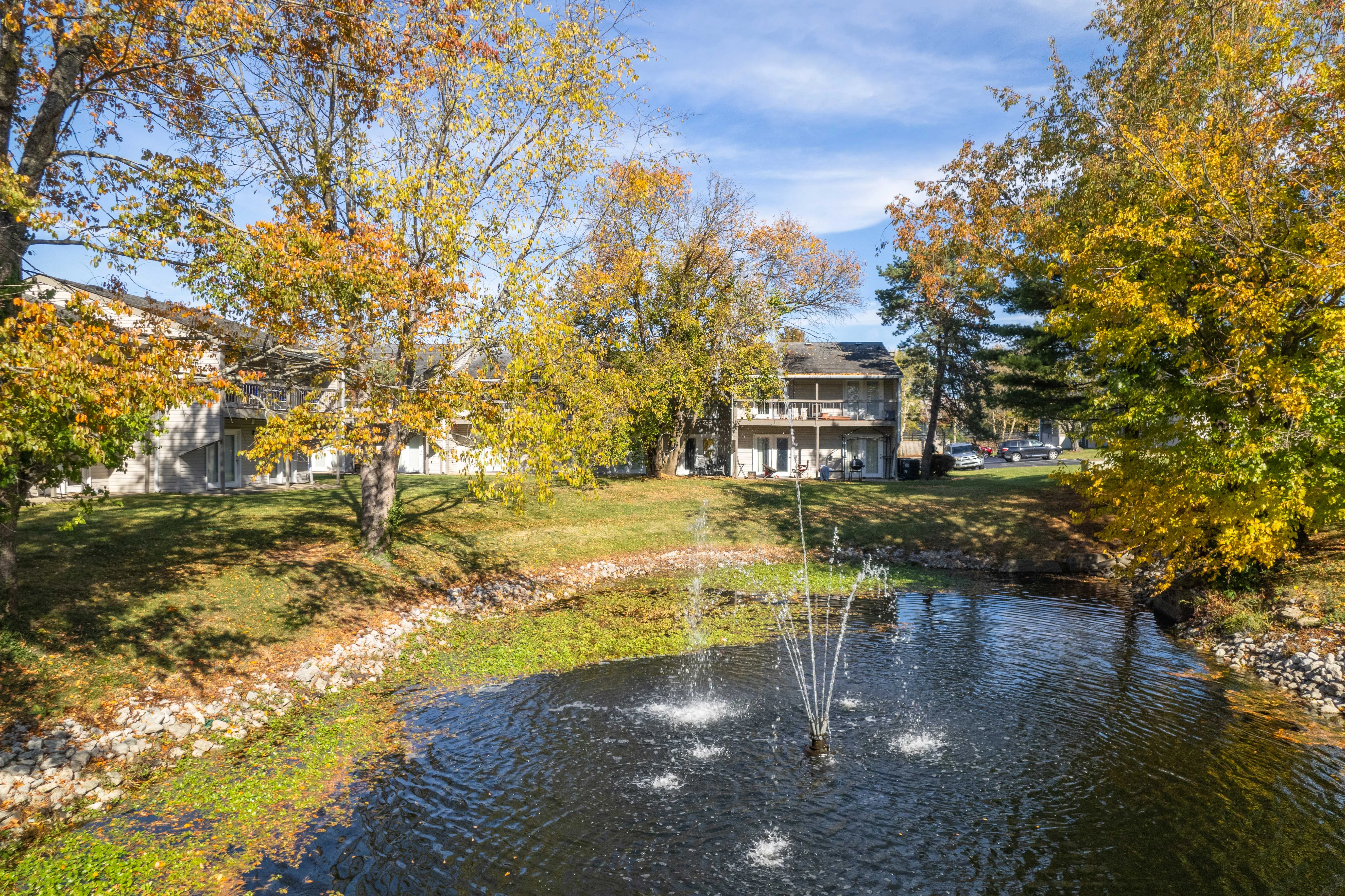 Autumn Pond with Fountain A scenic view of a pond with a fountain, surrounded by trees with autumn foliage and a building in the background.