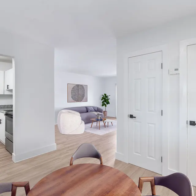 A modern apartment interior featuring a dining area with a round wooden table and stylish chairs. In the background, there's a kitchen with white cabinetry and a living room visible on the right, with a couch and a potted plant. Light wooden flooring enhances the bright atmosphere of the space.