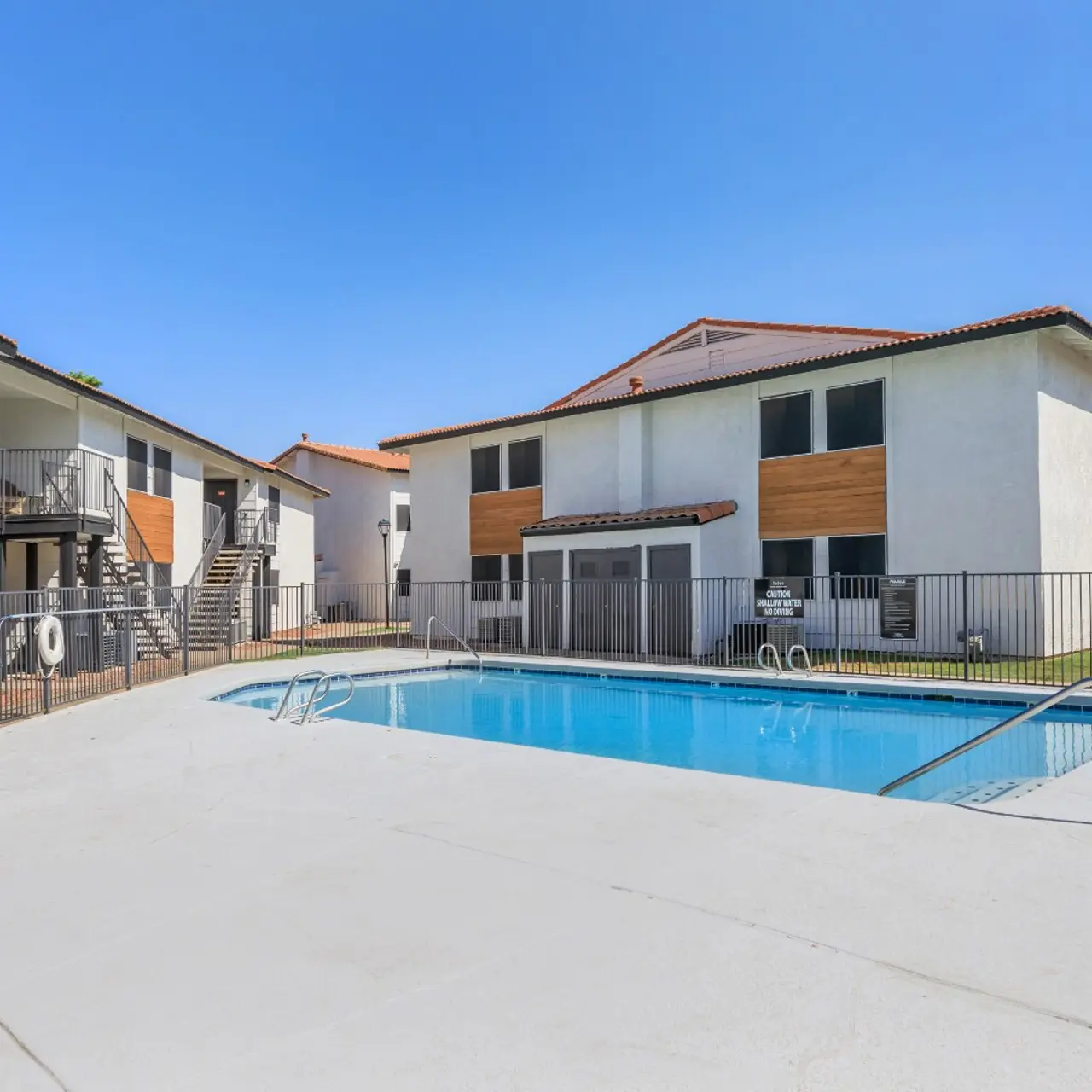 A view of a residential apartment complex featuring two buildings with balcony access and a swimming pool in the foreground. The pool area is surrounded by a fence and the sky is clear and sunny.