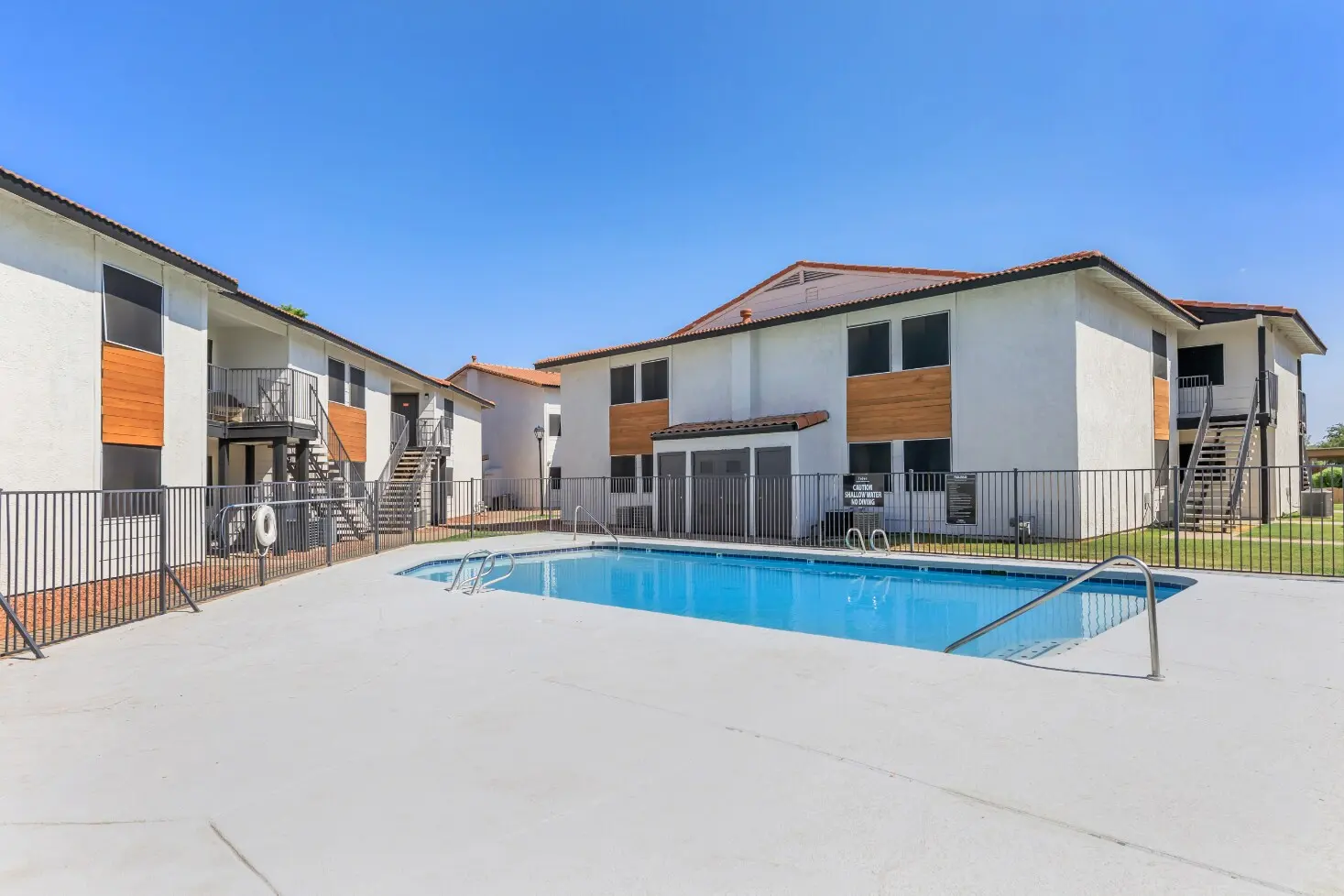 A view of a residential apartment complex featuring two buildings with balcony access and a swimming pool in the foreground. The pool area is surrounded by a fence and the sky is clear and sunny.