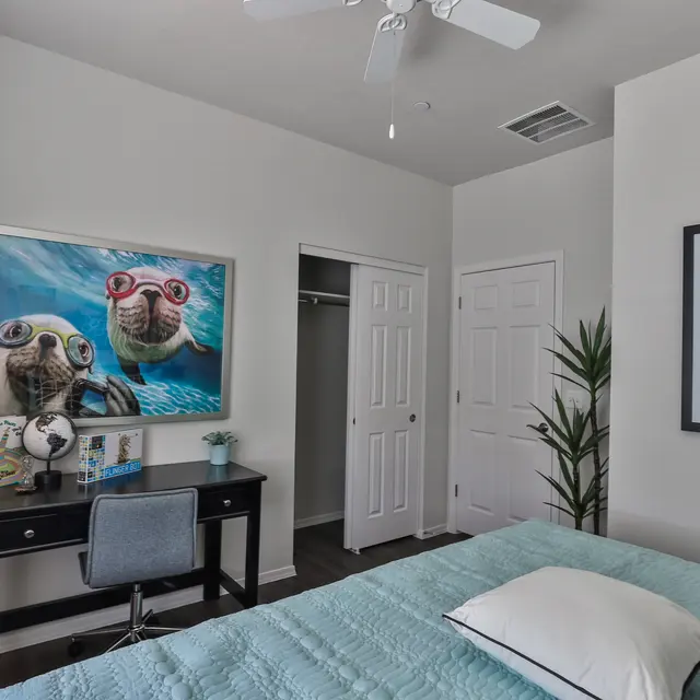 A modern bedroom featuring a light blue bedspread, a desk with a chair, a large framed picture of turtles wearing sunglasses on the wall, and a plant in the corner. Two doors can be seen leading to a closet and another room.