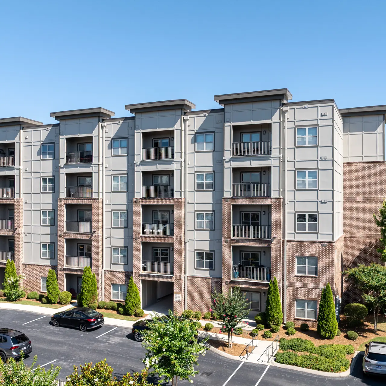 Modern Apartment Building A modern multi-story apartment building with a mix of brick and gray siding. The building has four floors with balconies on each level. Surrounding the building are landscaped areas with small trees and shrubs, along with a parking lot with multiple cars. The sky is clear and blue, indicating a sunny day.