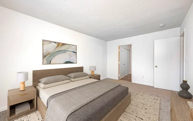 A modern bedroom featuring a bed with gray bedding, wooden nightstands, and decorative lamps. A large wall art piece is above the bed, and there are beige and cream-colored accents throughout the room.