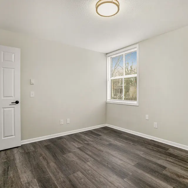 A vacant room with light gray walls and a wooden floor, featuring a window allowing natural light, a ceiling light fixture, and a closed white door.