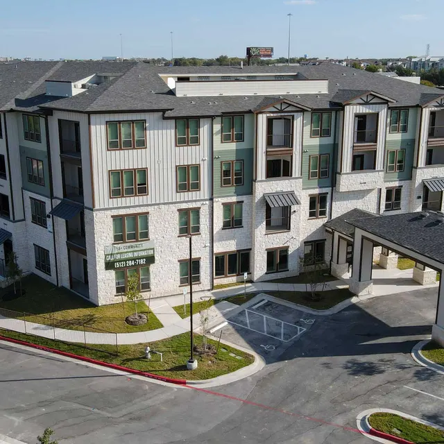 A modern apartment complex featuring multiple stories with a mixture of stone and siding exteriors. A paved parking area is visible alongside the building, with landscaped areas including small trees and grass. The entrance has a small covered awning.