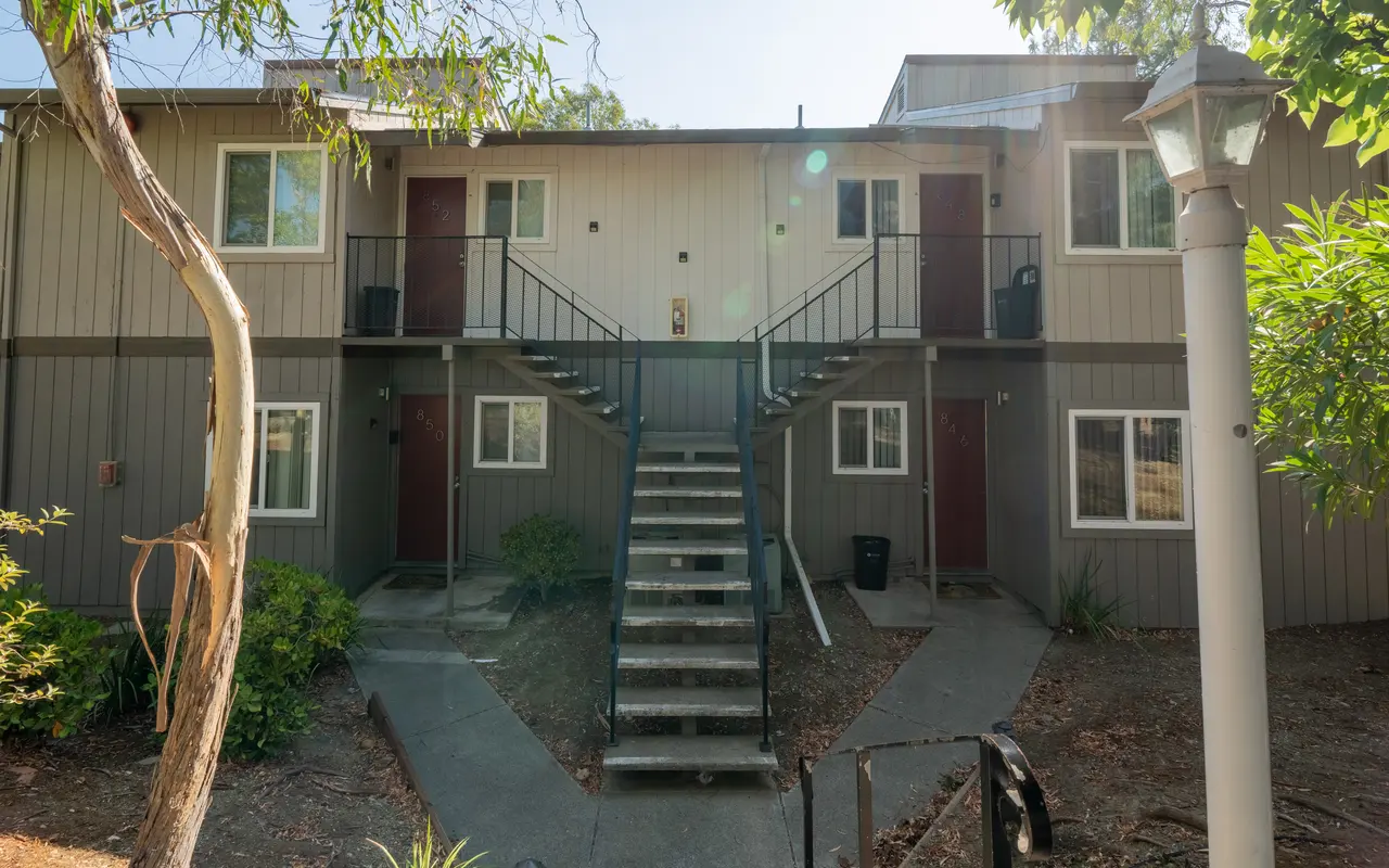 Two-story apartment building with stairs leading to the upper floor and green landscaping.