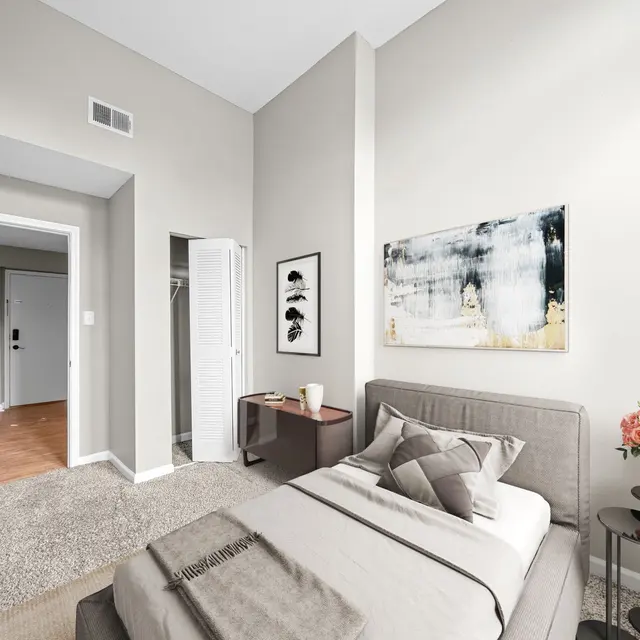 A modern bedroom featuring a single bed with neutral bedding, a decorative pillow, and a light gray wall. There is a wooden nightstand with a vase of flowers and a piece of abstract art on the wall. An open closet is visible, with a doorway leading to another room in the background.