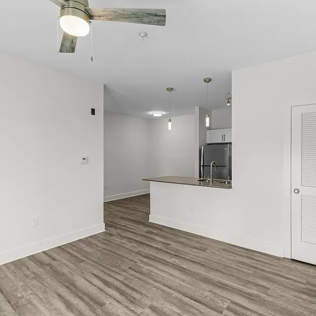 A modern apartment interior featuring an open layout with wood-style flooring, a ceiling fan, and a kitchen area visible in the background.