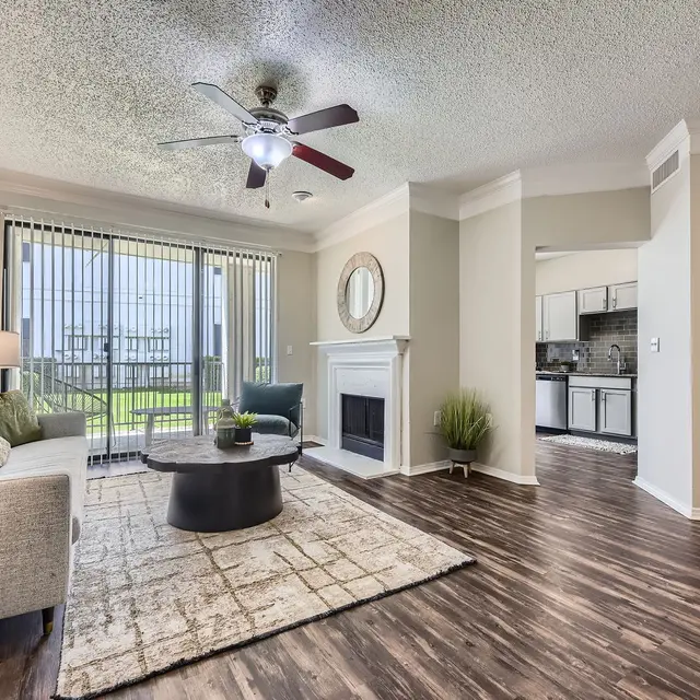 A stylish living room featuring a light-colored sofa, a circular coffee table, and a rug on hardwood flooring. There is a ceiling fan and large windows with blinds allowing natural light. A fireplace is present on one wall, and the space has a modern aesthetic.