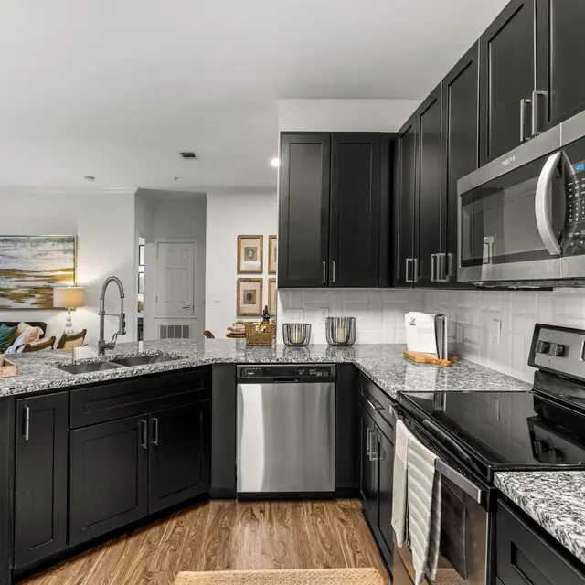 A modern kitchen featuring black cabinets and granite countertops, stainless steel appliances, and a spacious layout with an adjacent seating area visible.