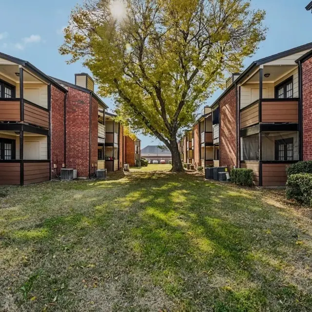 A view of an apartment complex with two buildings on either side of a grassy area. A large tree stands in the middle, casting shadows on the ground. The buildings are red brick with brown accents and balconies.